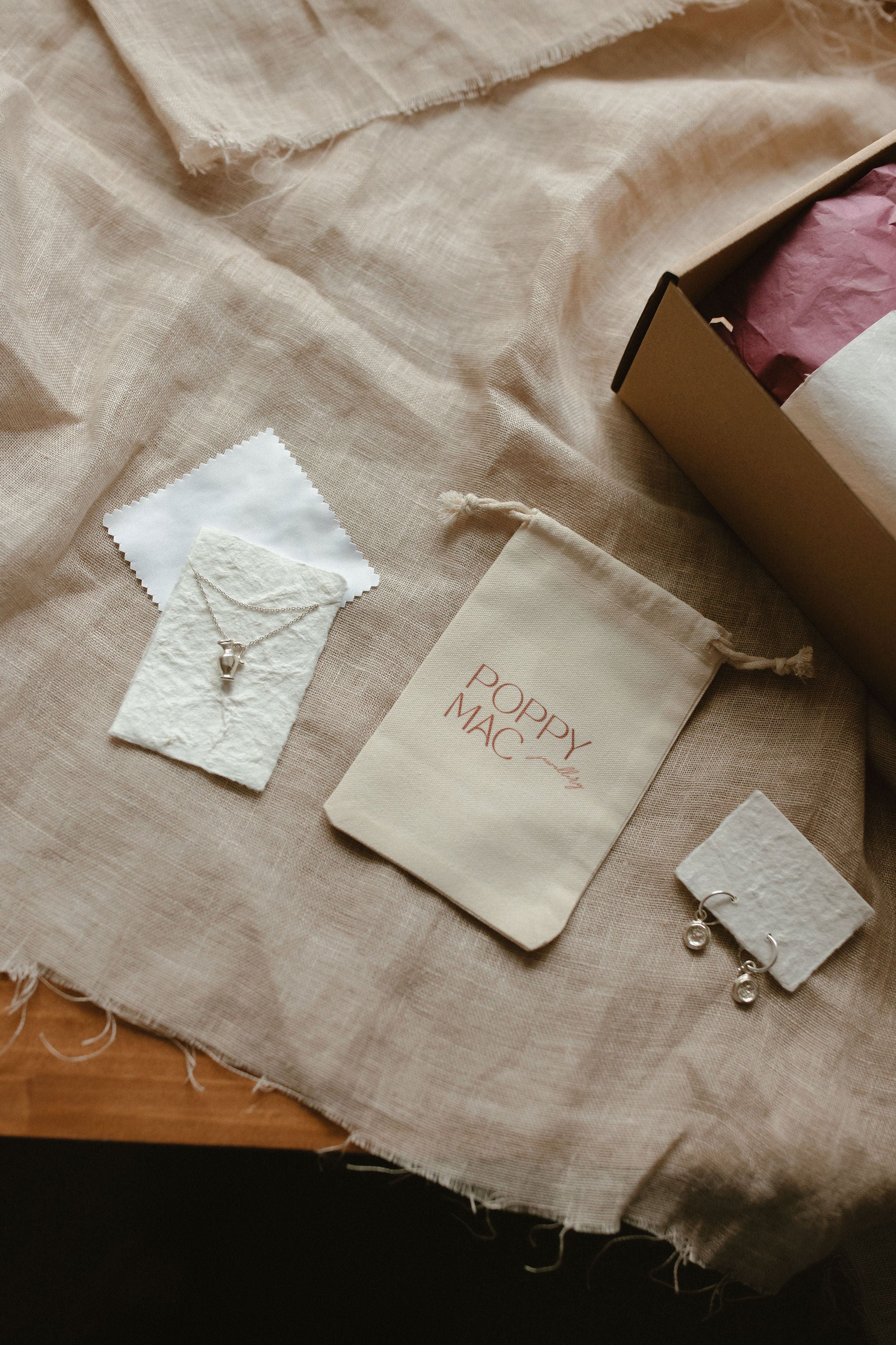 A styled flat-lay of jewelry packaging and accessories on a beige linen fabric background, including a cream drawstring pouch with pink 'Poppy Mac' embroidery, small white textured cards, and a cardboard box corner.