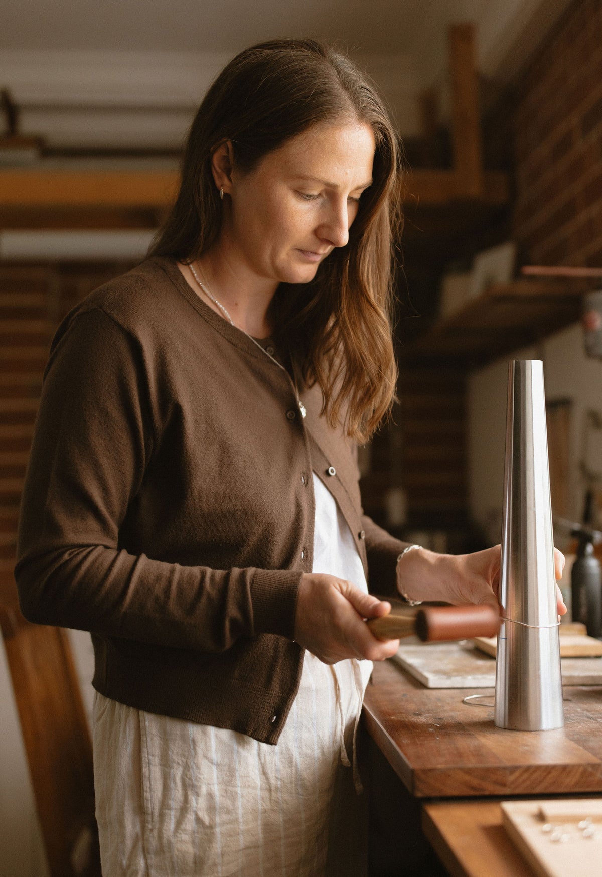 A woman in a brown cardigan and white dress stands at a wooden workbench, holding a metal object next to a tall silver cone-shaped tool in a warm, rustic workshop.