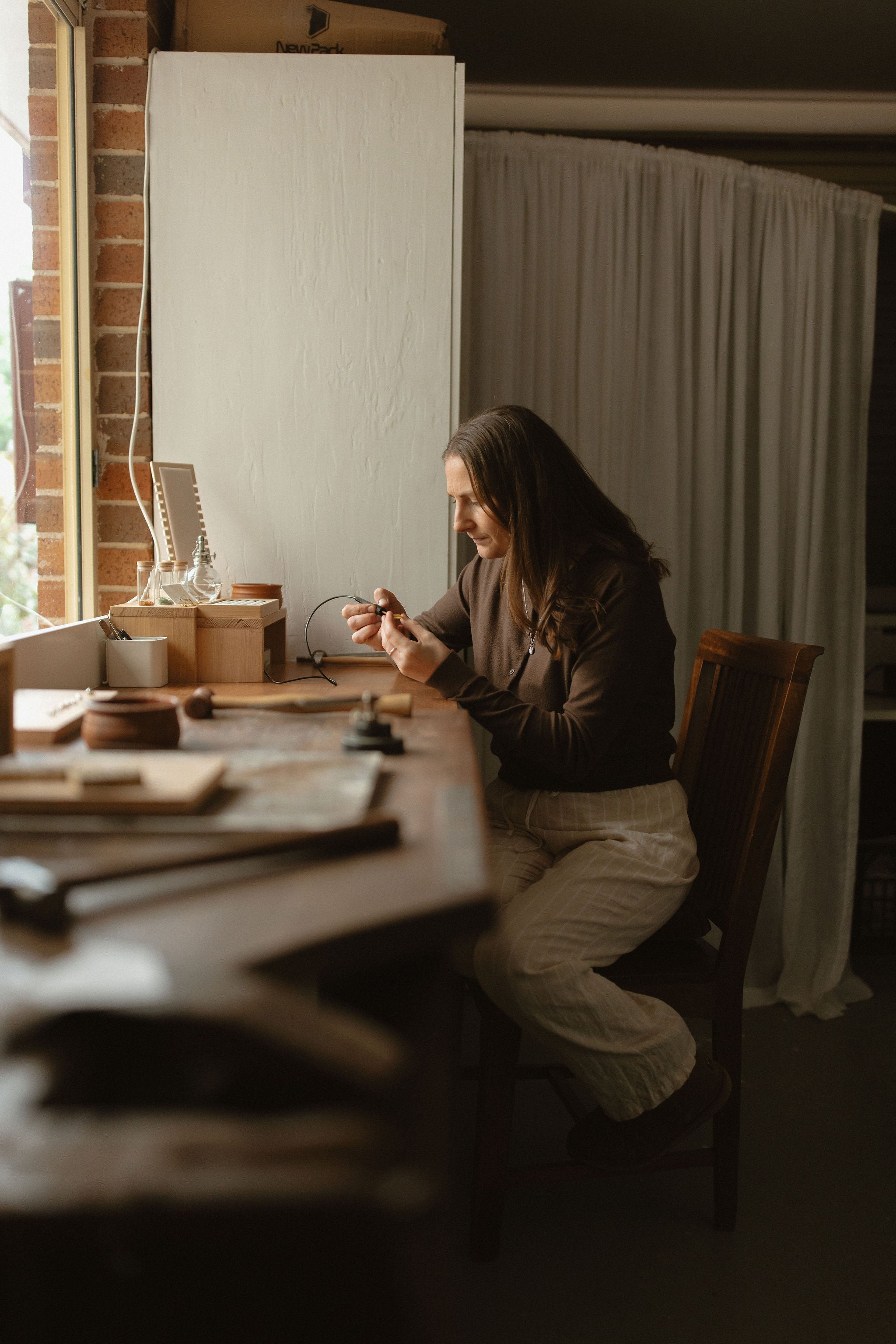 A woman sits at a wooden table in a warm, rustic room, working on a small craft or writing instrument. The scene is softly lit with natural light from a window, featuring a whiteboard, brick wall, and draped curtains in the background.